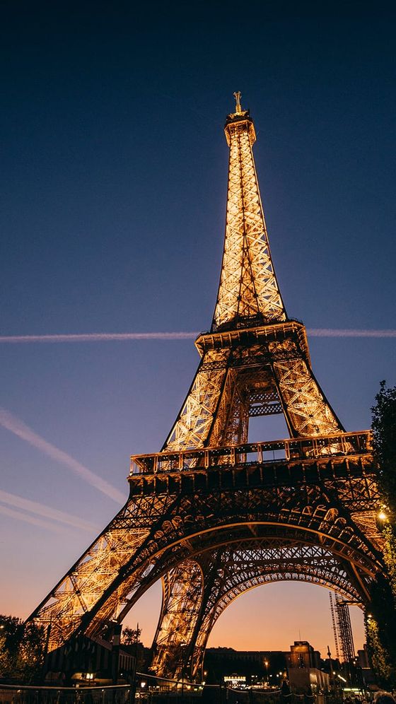 Lit-up Eiffel Tower at dusk, against a deep blue sky with airplane trails, surrounded by silhouettes of trees and buildings.