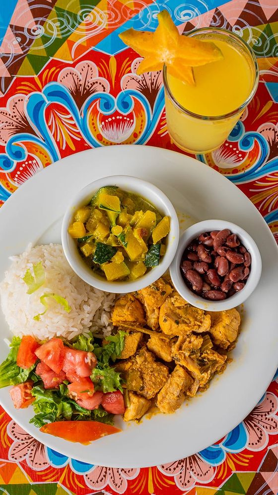 A vibrant meal with rice, curry, beans, salad, and grilled chicken on a patterned tablecloth; a starfruit-topped drink is alongside.