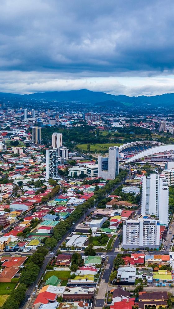Aerial view of a cityscape with colorful houses, tall buildings, and a stadium, set against a backdrop of green hills and a cloudy sky.