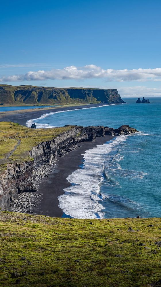 Coastal scene with cliffs, a black sand beach, and ocean waves. Green landscape and clear blue sky in the background.