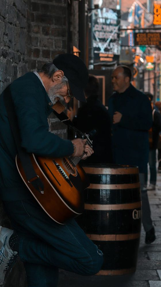 Bearded man in a cap plays guitar while leaning against a brick wall in a dimly lit alley, with people walking by and neon signs in the background.