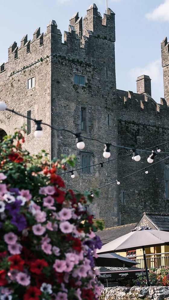 Stone castle with crenelations against a blue sky. Colorful flowers and string lights are in the foreground, adding a festive touch.
