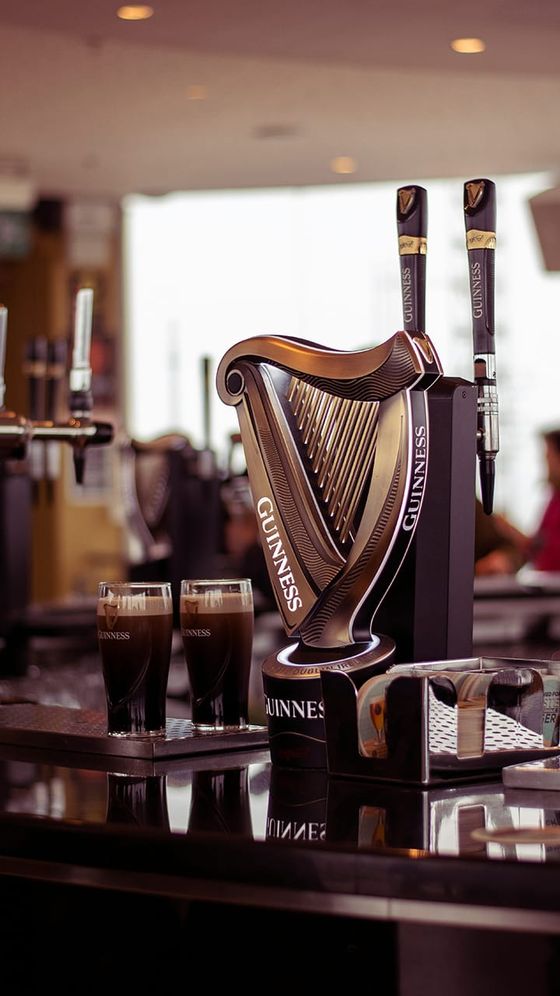 Two pints of Guinness stout sit on a glossy bar next to a Guinness harp tap handle, with a blurred background.