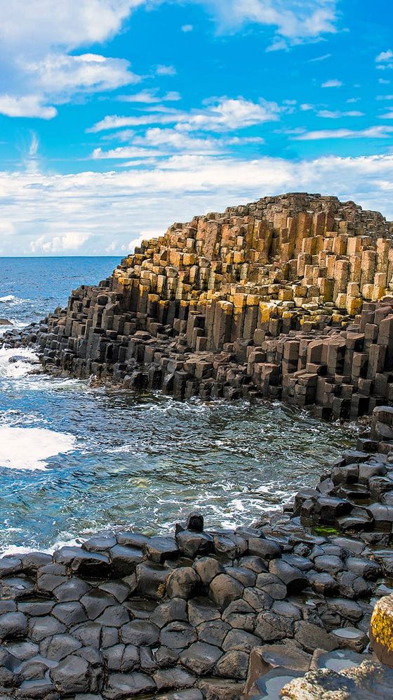 Hexagonal rock columns rise from the ocean at Giant's Causeway, under a bright blue sky with scattered clouds.