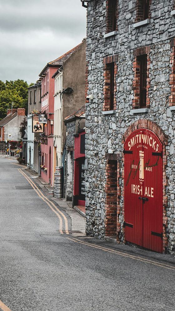 A quaint street with stone buildings, a large red door with "Smithwick's Irish Ale," and colorful facades under a cloudy sky.