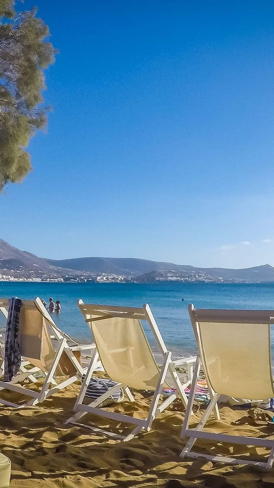 Beach scene with empty lounge chairs on sandy shore, clear blue sea, and distant hills under a bright blue sky.
