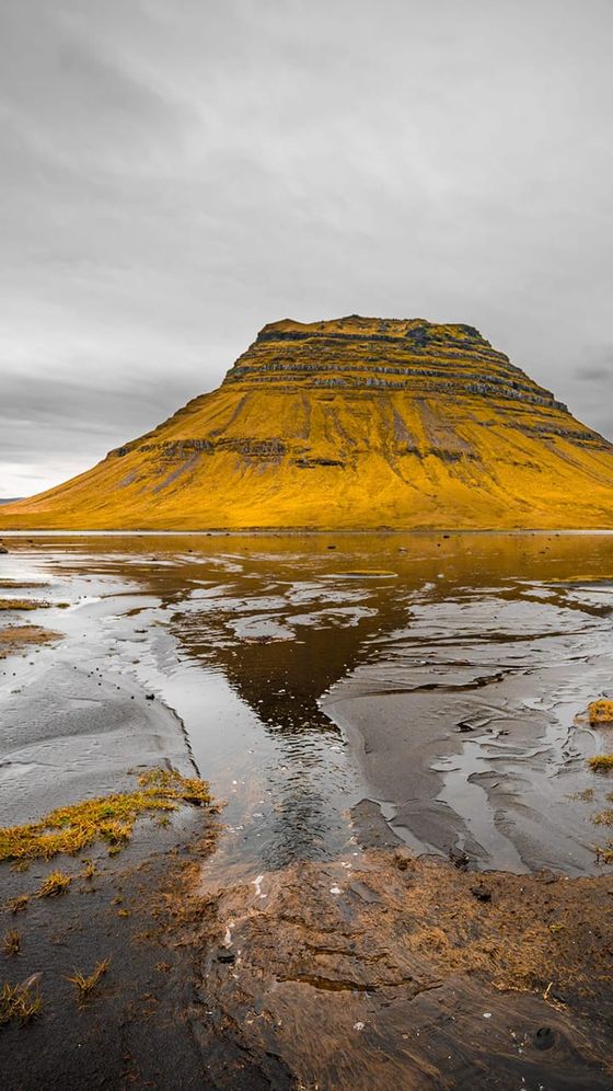Mountainside with layered rock formations, surrounded by golden-brown fields and reflecting in a shallow, rocky pond under a cloudy sky.