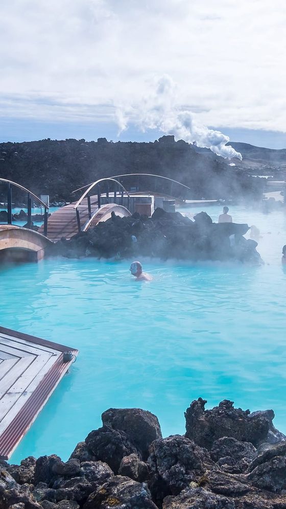 People relaxing in the steamy blue waters of Iceland's Blue Lagoon, surrounded by rocky terrain and a wooden bridge under a cloudy sky.