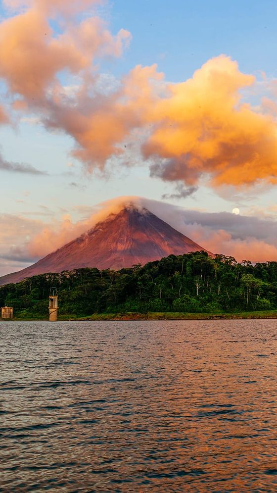 A scenic view of a conical volcano with a pink and orange sky at sunset, surrounded by lush greenery and a calm lake in the foreground.