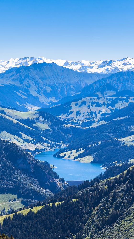 Scenic view of a mountainous landscape with a blue lake, green valleys, and distant snow-capped peaks under a clear blue sky.