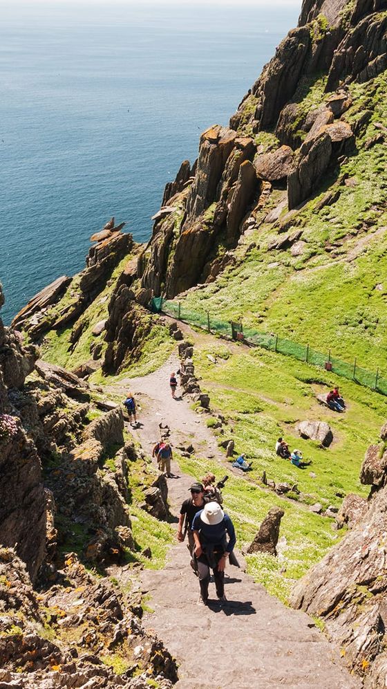 People climbing a steep stone staircase on a grassy, rocky hill with a view of the ocean in the background.