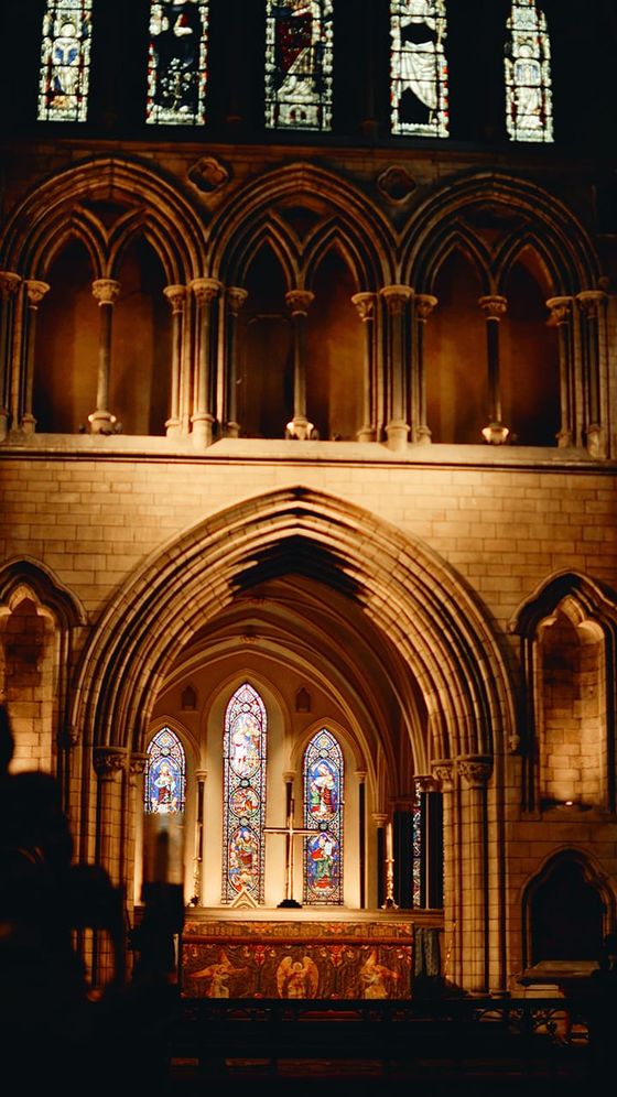 A dimly lit cathedral interior with arched stone architecture and vibrant stained glass windows above an ornate altar.