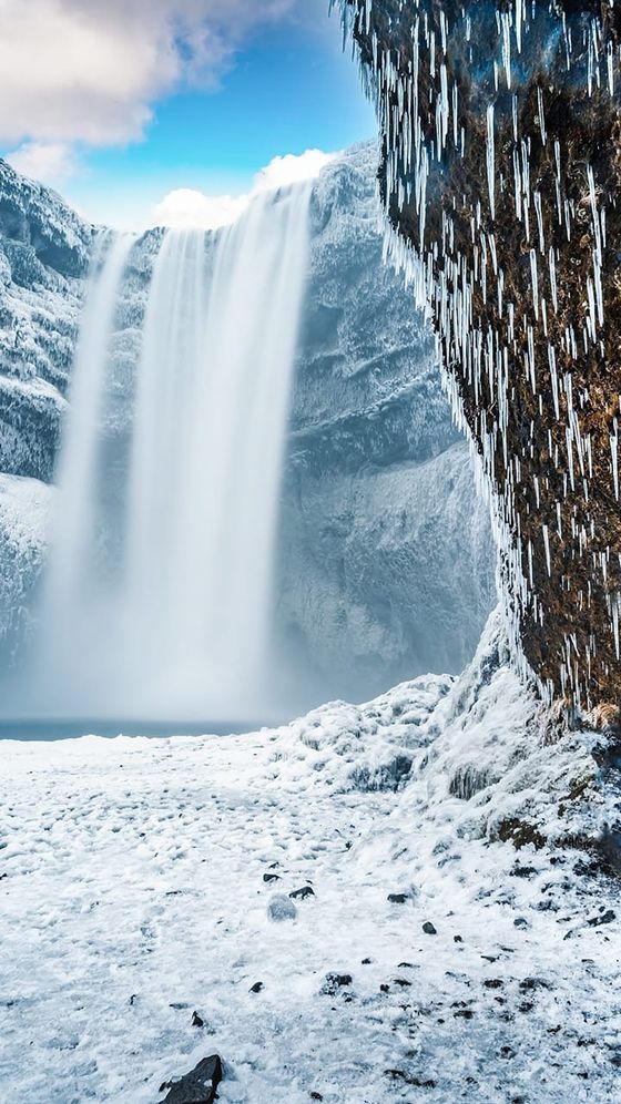Frozen waterfall with icicles hanging from a cliff, surrounded by snow and ice, under a bright blue sky.