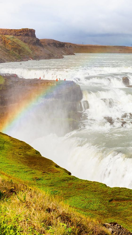 A powerful waterfall cascades over rocky cliffs, surrounded by lush green terrain, with a vivid rainbow arching through the mist.