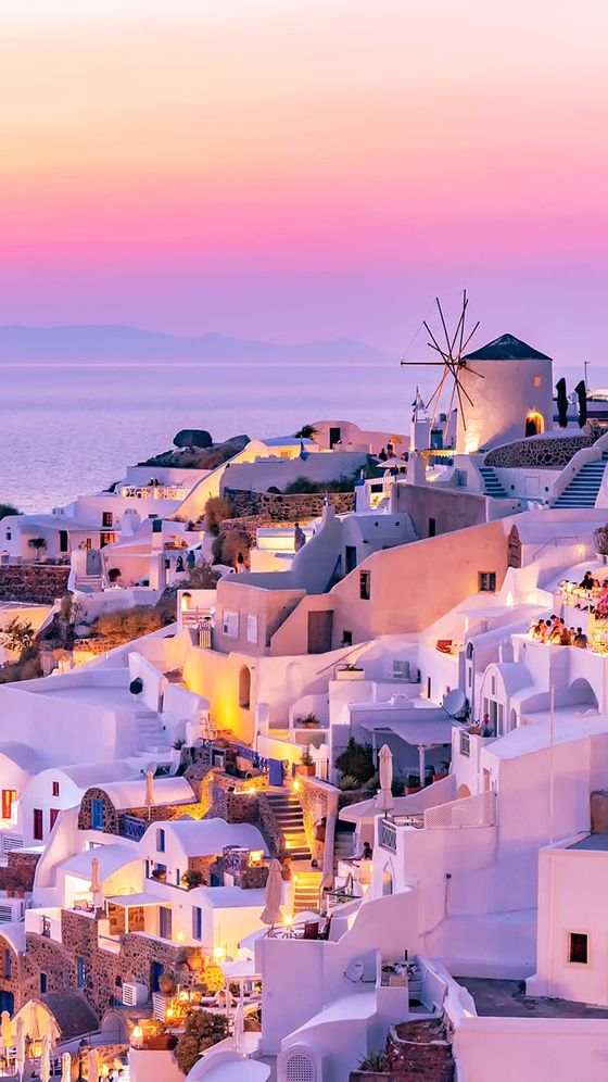 Santorini at sunset with whitewashed buildings, a windmill, and a pink-orange sky overlooking the sea.