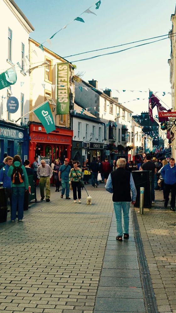 A bustling street scene in a lively town, with people walking past colorful storefronts and flags hanging above in the sunny weather.