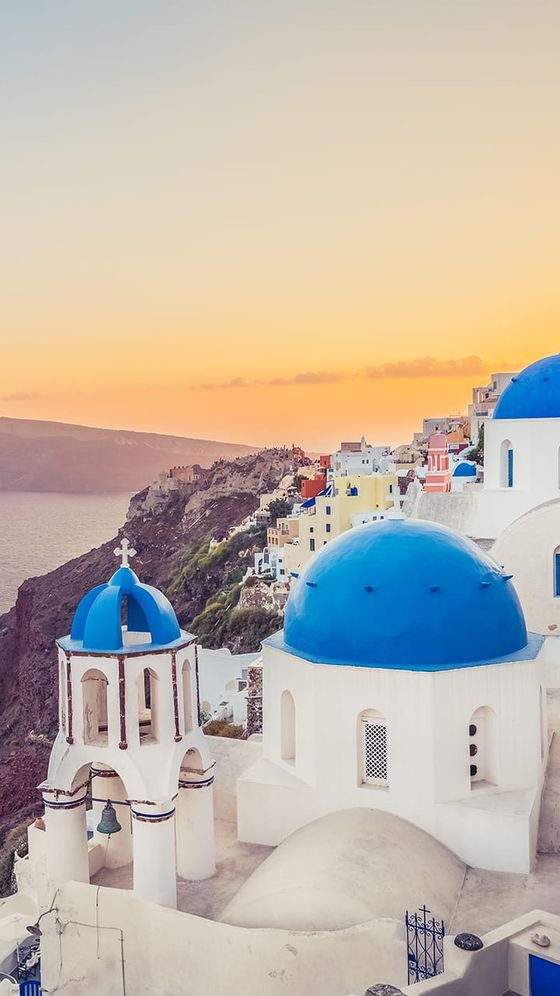 Scenic view of Santorini with iconic blue-domed churches and white buildings overlooking a sunset-lit sea and rugged coastline.