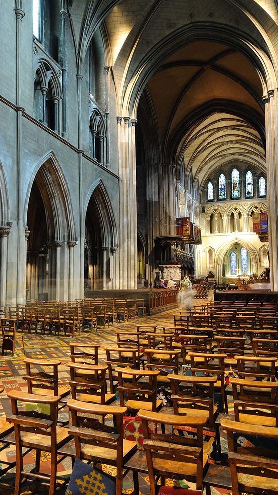 Interior of a grand cathedral with tall arches, stained glass windows, wooden chairs, and a patterned carpet under a high vaulted ceiling.
