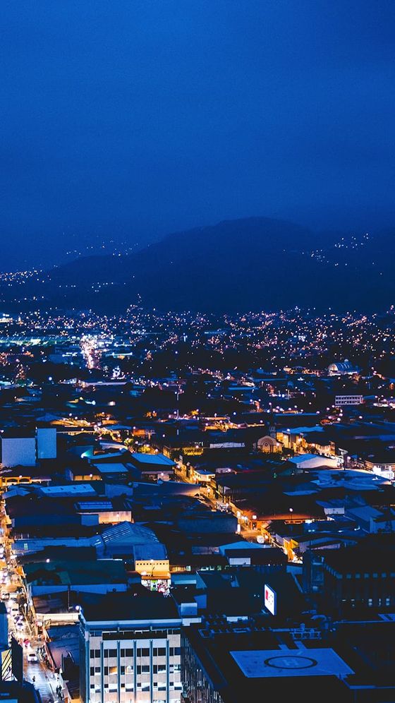 Cityscape at night with lit streets and buildings, distant hills, and a deep blue sky, creating a serene urban atmosphere.