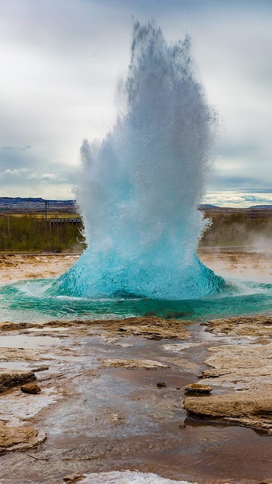 A vibrant geyser erupts with a powerful surge of blue water under a cloudy sky, surrounded by rocky terrain.