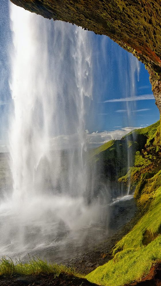Waterfall cascading into a pool, surrounded by lush green cliffs under a blue sky, viewed from behind the falling water.