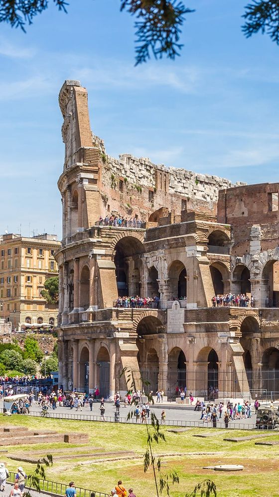 The Colosseum in Rome on a sunny day, with tourists exploring its ancient, partly-ruined structure, and people walking in the foreground.