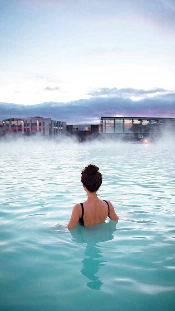 A person in a black swimsuit stands in a steaming blue lagoon, facing modern buildings under a cloudy sky.