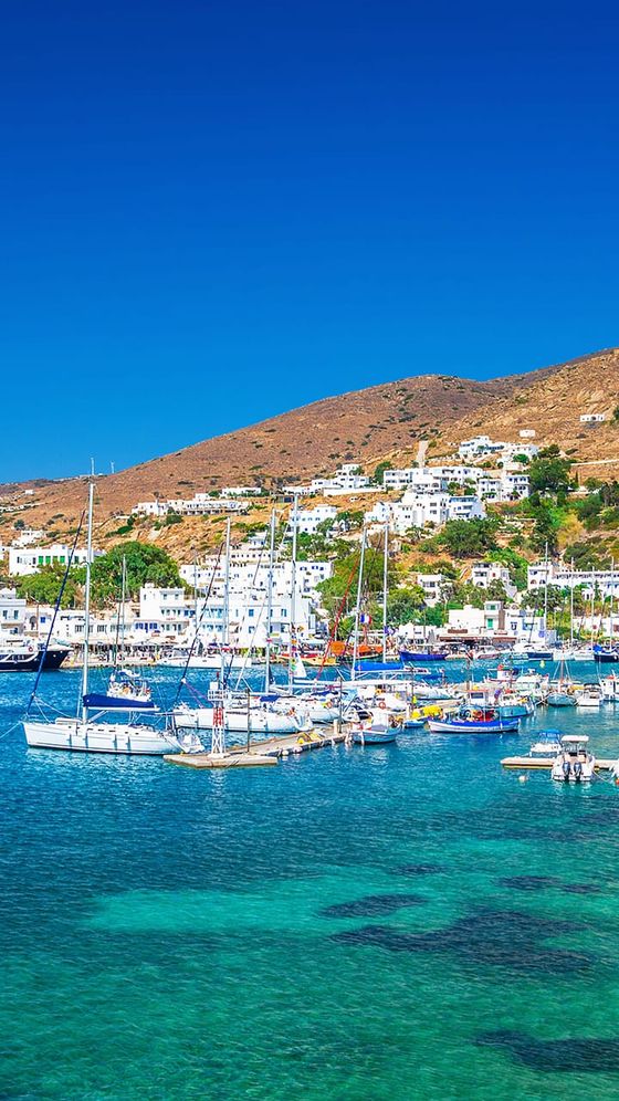 A scenic coastal town with white buildings on a hillside, clear blue water, and sailing boats in a marina under a bright blue sky.