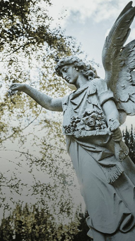 Statue of an angel with outstretched arm holding flowers, surrounded by trees and a cloudy sky in the background.