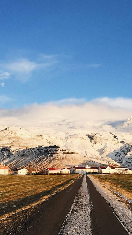 A narrow road leads to a settlement with red-roofed buildings, set against a backdrop of snow-covered mountains under a clear blue sky.
