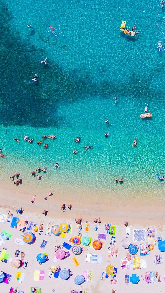 Aerial view of a sunny beach with colorful umbrellas and beachgoers on the sand, and people swimming in the clear turquoise sea.