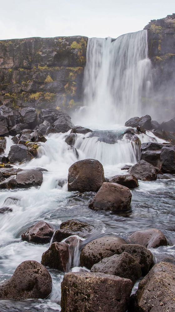 A waterfall cascades over a rocky cliff into a stream, surrounded by moss-covered stones and large boulders.
