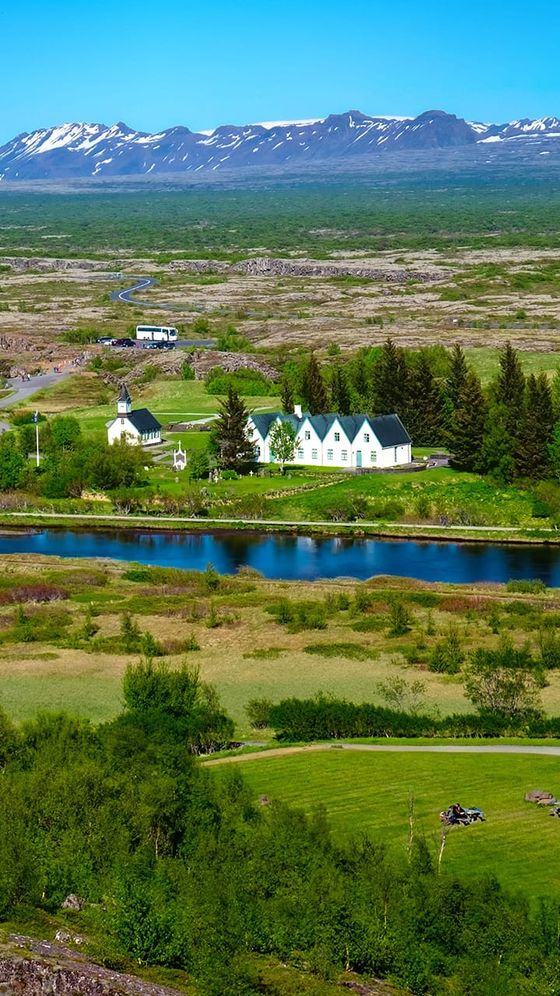 Scenic landscape with a small white church and houses by a river, surrounded by lush greenery, with distant snow-capped mountains under a clear blue sky.