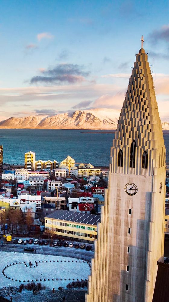Aerial view of Reykjavik with the Hallgrímskirkja church in the foreground, snowy mountains, and ocean in the background.