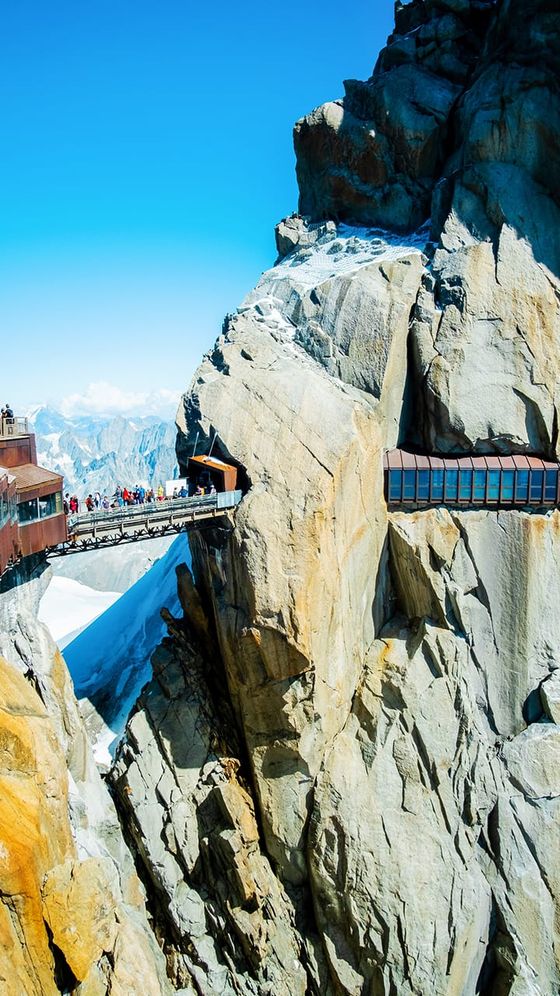 A narrow pedestrian bridge connects two rocky mountain peaks under a clear blue sky, with people walking along it. Snow is visible on the peaks.