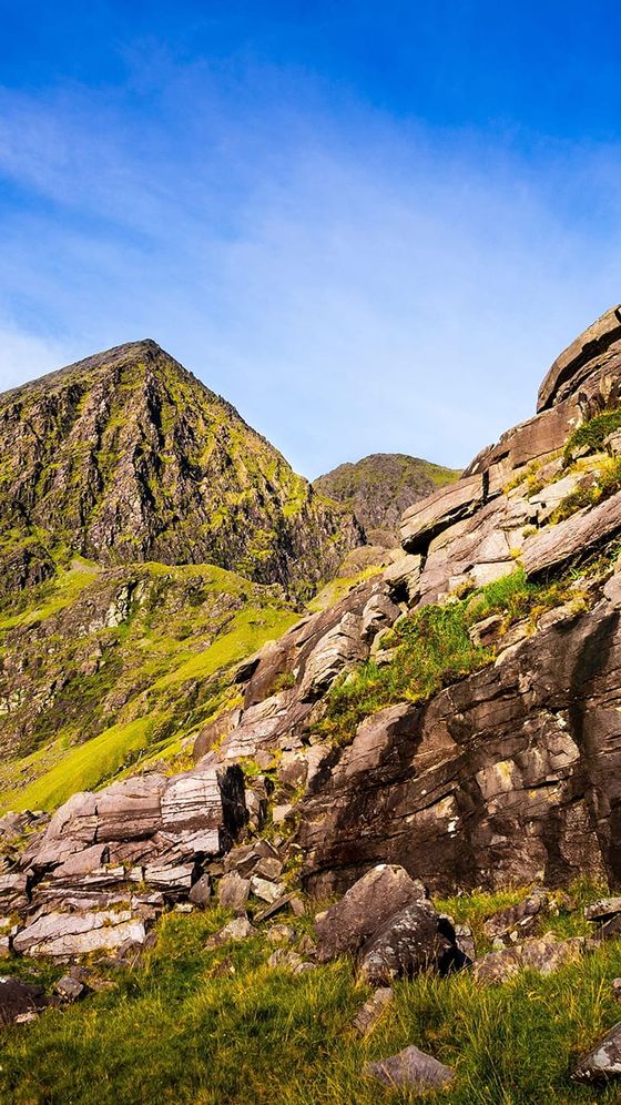 Rocky mountain landscape with green slopes under a clear blue sky, featuring rugged terrain and scattered boulders.