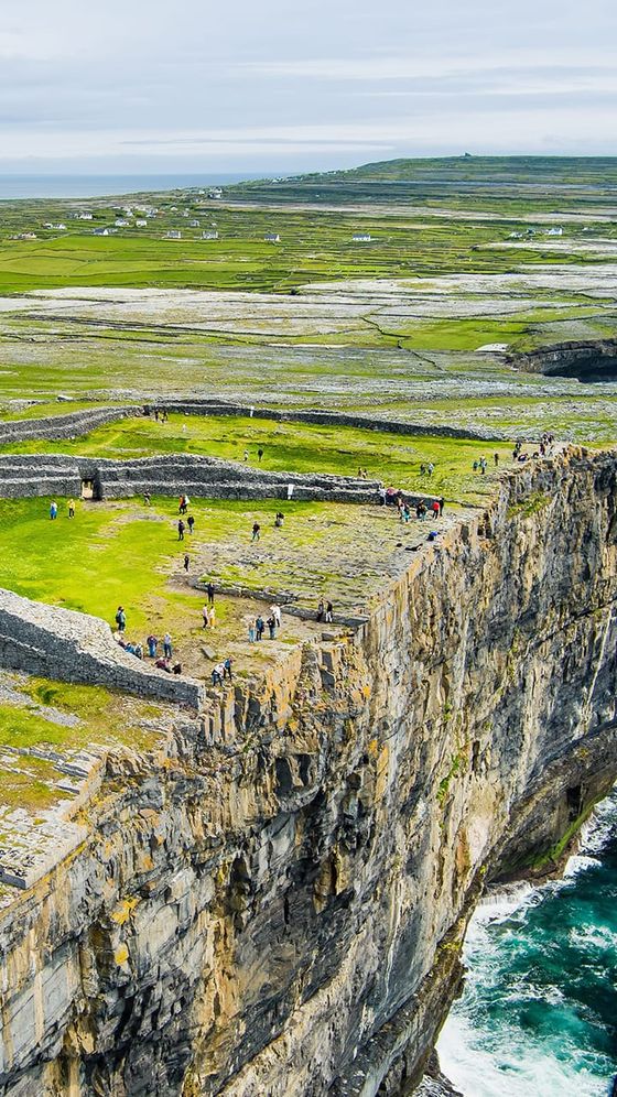 Aerial view of people visiting Inis Mór's cliffside fort, featuring lush green fields and rocky cliffs above the ocean.