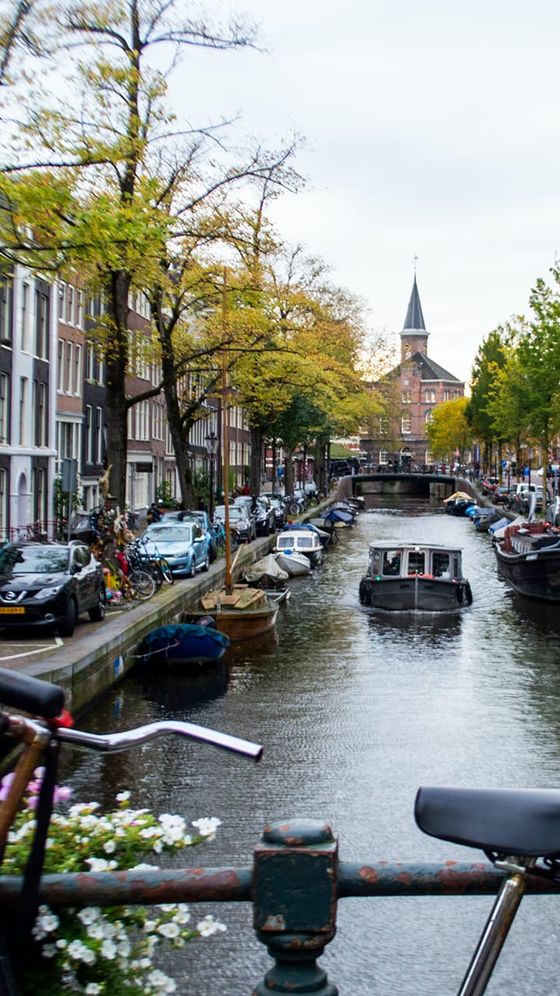 Charming canal scene in Amsterdam with boats, parked cars, bicycles, and historic brick buildings lining the waterfront, trees in autumn.