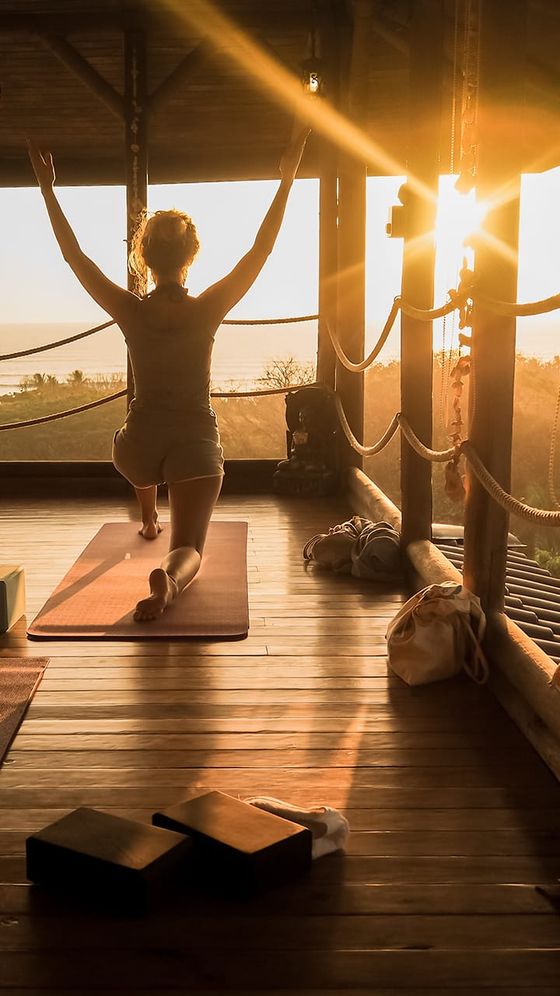 A person practices yoga on a mat in a serene, sunlit room with wooden floors and large windows, facing the sunrise.