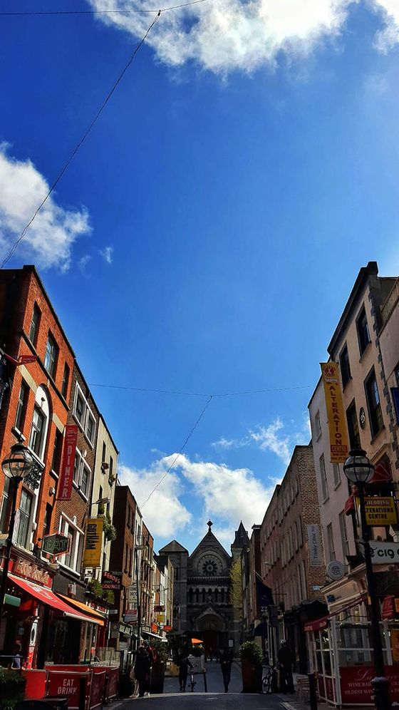 A narrow street bustling with people, flanked by colorful buildings and signs, leads to a distant church under a bright blue sky.