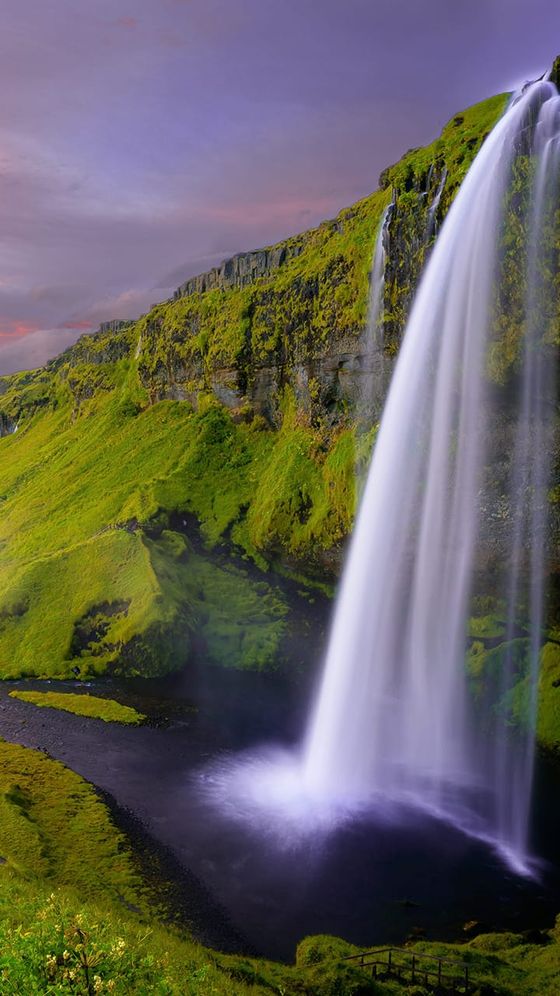 A tall waterfall cascading down a lush green cliff into a serene pool, under a purple-tinged sky at sunset.