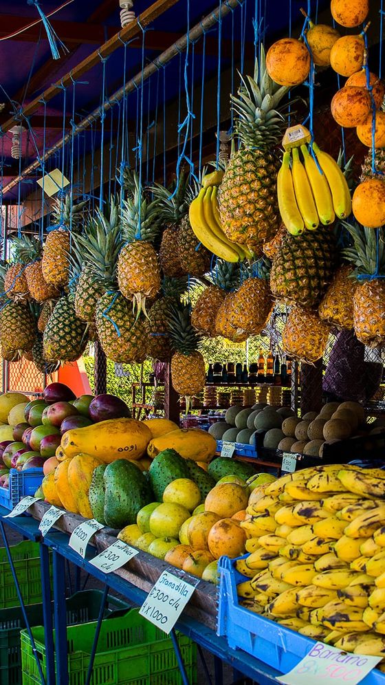 Colorful fruit market display with hanging pineapples, bananas, mangoes, and other tropical fruits stacked in rows.
