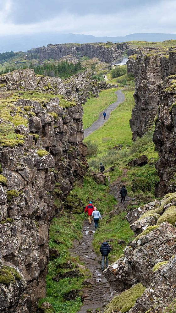 People walking a rocky path between moss-covered cliffs in a lush, green landscape under a cloudy sky.