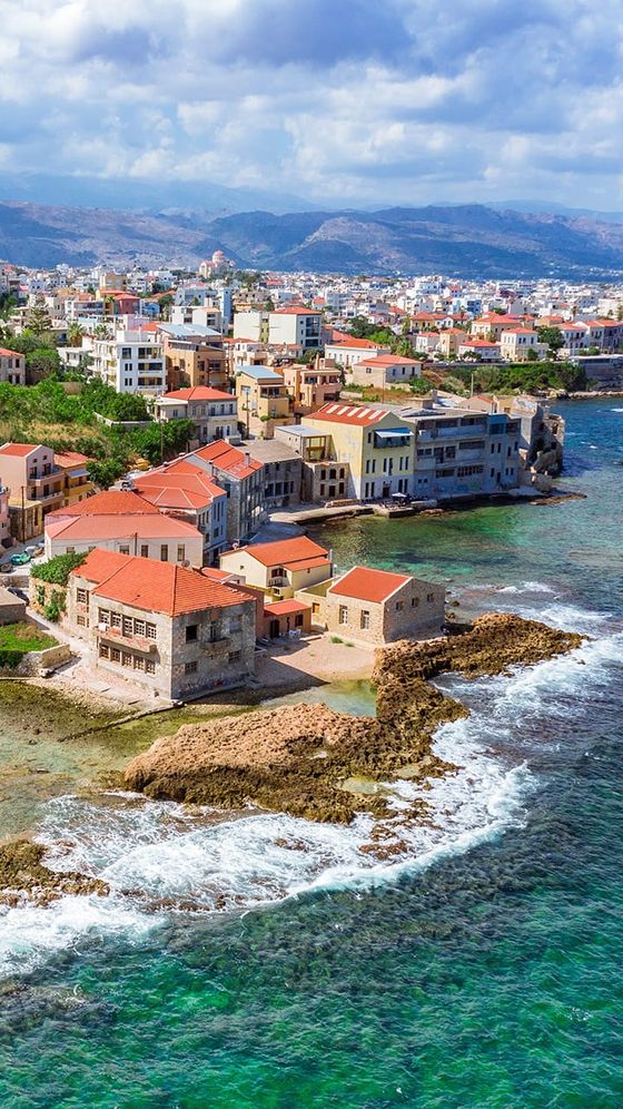 Aerial view of a coastal town with colorful buildings, rocky shoreline, and clear turquoise water under a partly cloudy sky.