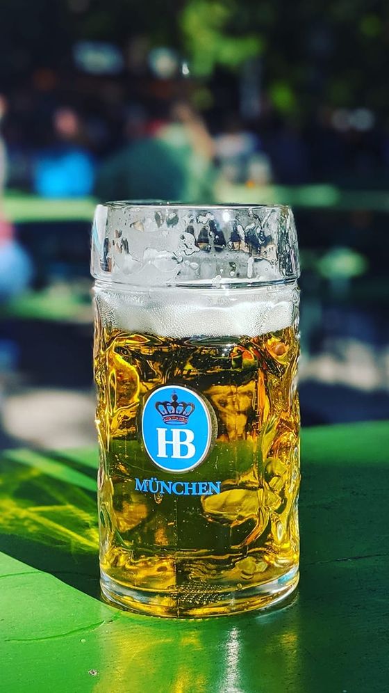 A glass stein with blonde beer and foam, branded "München HB," sits on a green table outdoors in dappled sunlight.