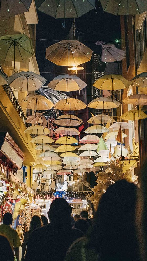 A bustling alley at night, with numerous umbrellas hanging above, lit shops on either side, and people walking beneath.