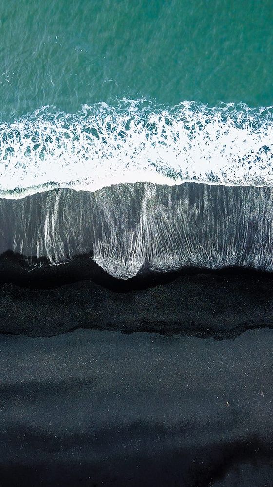 Aerial view of waves gently crashing onto a black sand beach with turquoise water, creating a striking contrast between sea and shore.