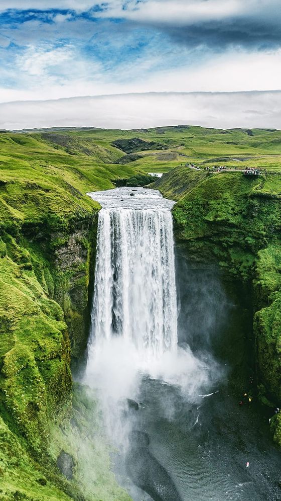 Waterfall cascading down a lush green cliffside into a river below, with a cloudy sky above and people visible on the horizon.