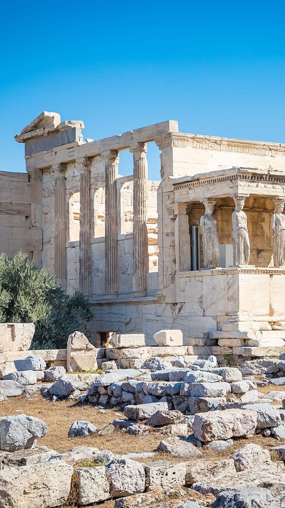 Ancient Greek temple ruins with stone columns and caryatids under a clear blue sky, surrounded by scattered stones and sparse vegetation.