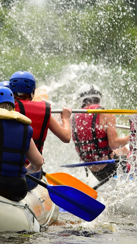 People rafting in turbulent water, wearing helmets and life vests, paddling vigorously as water splashes around them.